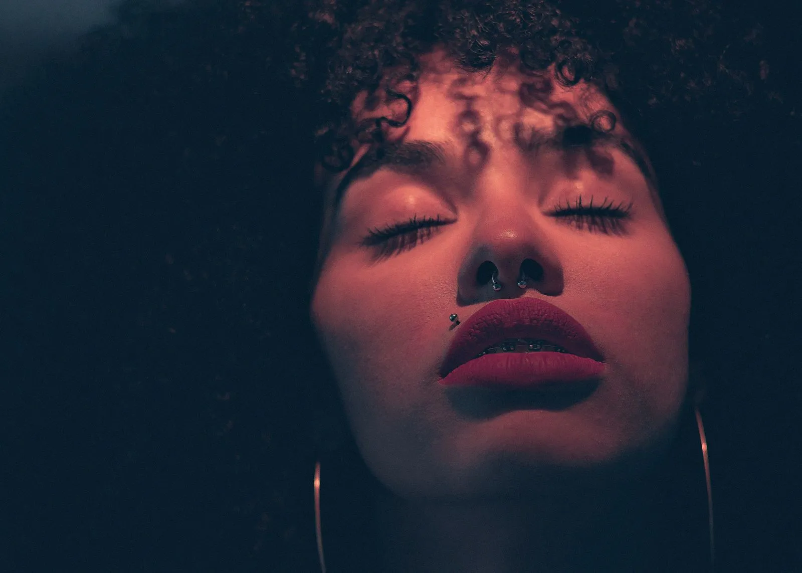 A dramatic close-up portrait of a woman with curly hair and nose piercings.